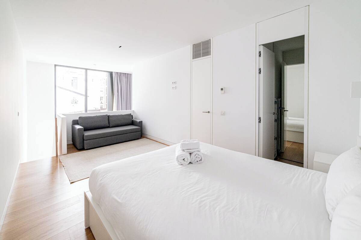 Una habitación de hotel moderna con una cama grande, un sofá gris y una ventana con cortinas.