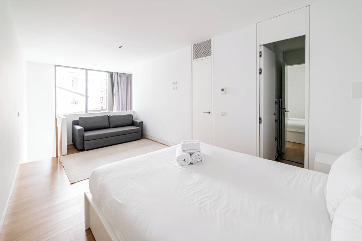 A modern hotel room with a large bed, a grey sofa, and a window with curtains.
