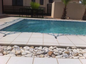 Una piscina con una cubierta de red azul, rodeada de baldosas blancas y un borde de guijarros.