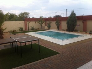 Una piscina rectangular con borde de azulejos, rodeada por un patio de ladrillo y césped artificial verde.