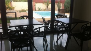 Un patio con mesas y sillas, piso de baldosas y vista a una piscina y follaje al fondo.