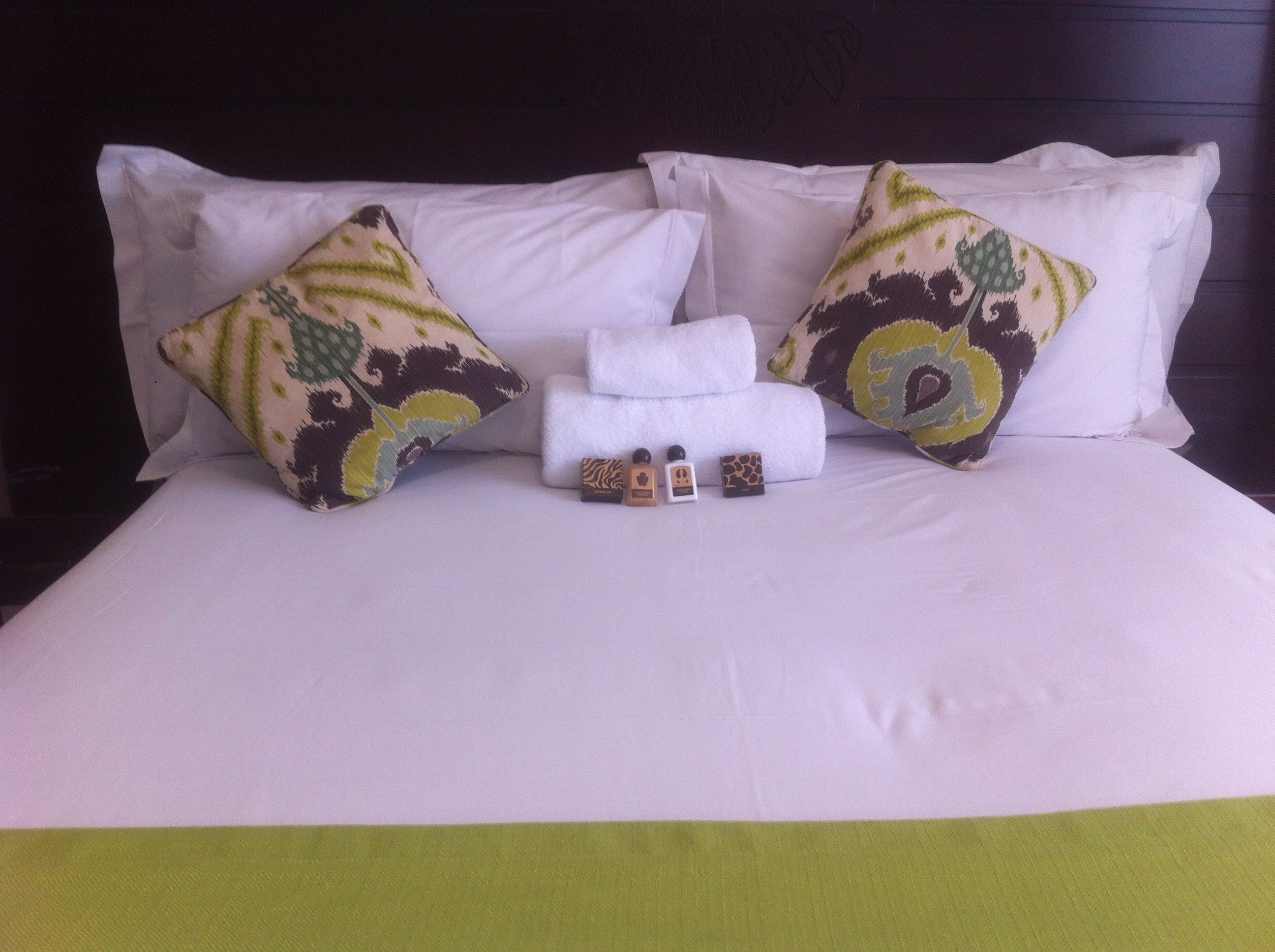 Un lit bien fait, avec du linge de lit blanc et des coussins décoratifs, une pile de serviettes pliées et une petite bouteille de lotion.
