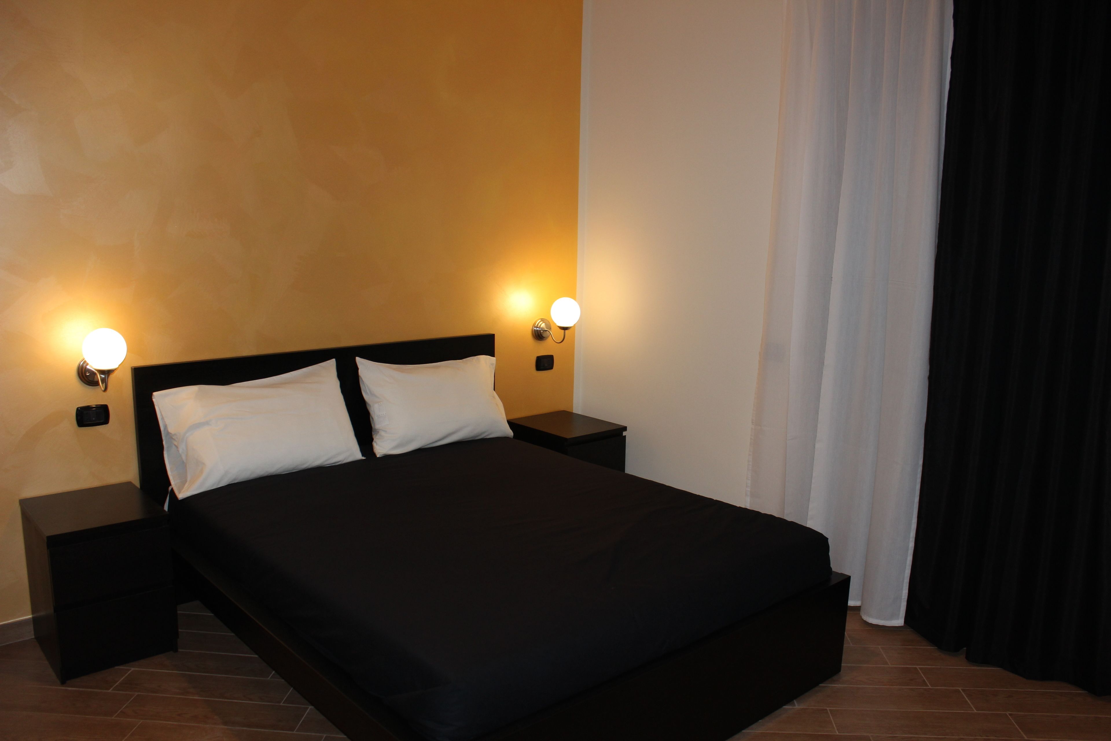 A neatly made bed with a black bedspread and white pillows, flanked by two bedside tables with wall-mounted lamps.