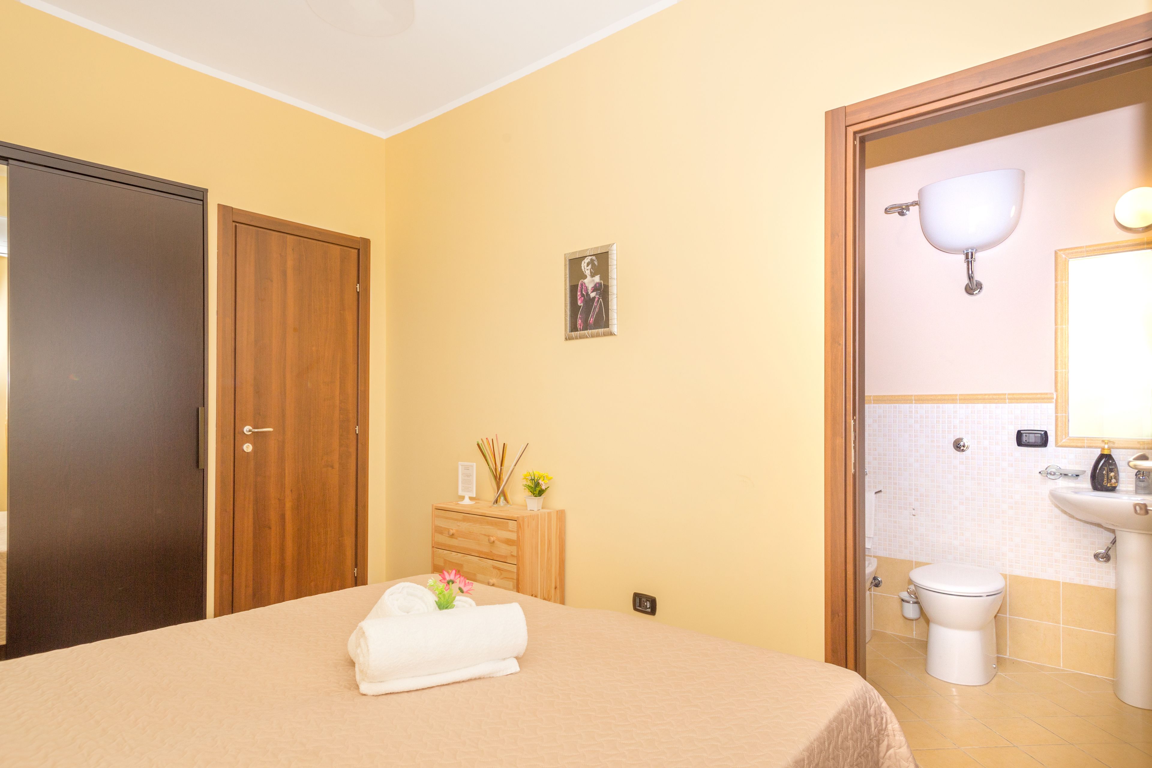 Une chambre d’hôtel comprenant un lit, une armoire en bois, une petite commode en bois avec des articles de toilette, un tableau encadré au mur, et une salle de bain avec un lavabo et des toilettes.