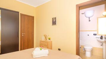 Une chambre d’hôtel comprenant un lit, une armoire en bois, une petite commode en bois avec des articles de toilette, un tableau encadré au mur, et une salle de bain avec un lavabo et des toilettes.