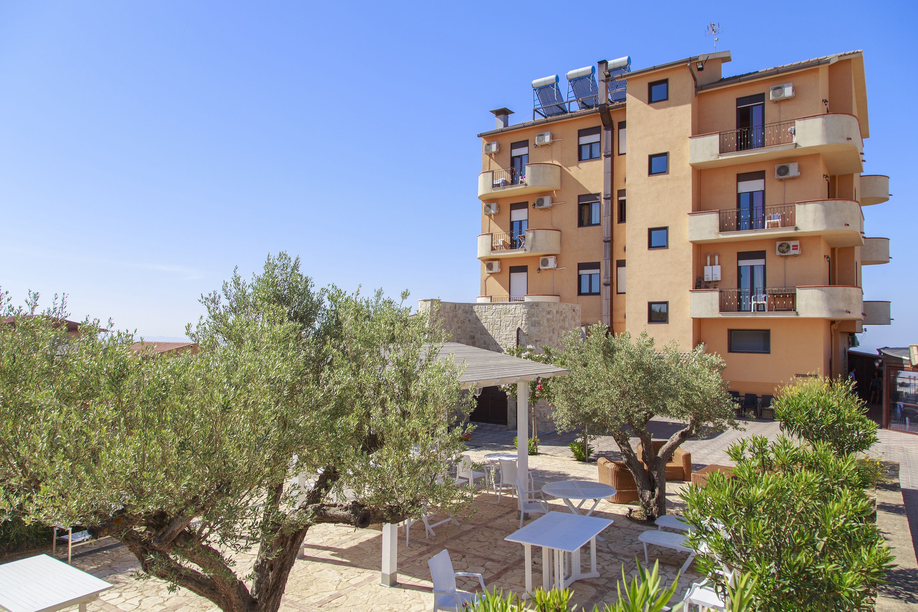 A multi-story building with balconies, outdoor seating, and olive trees.