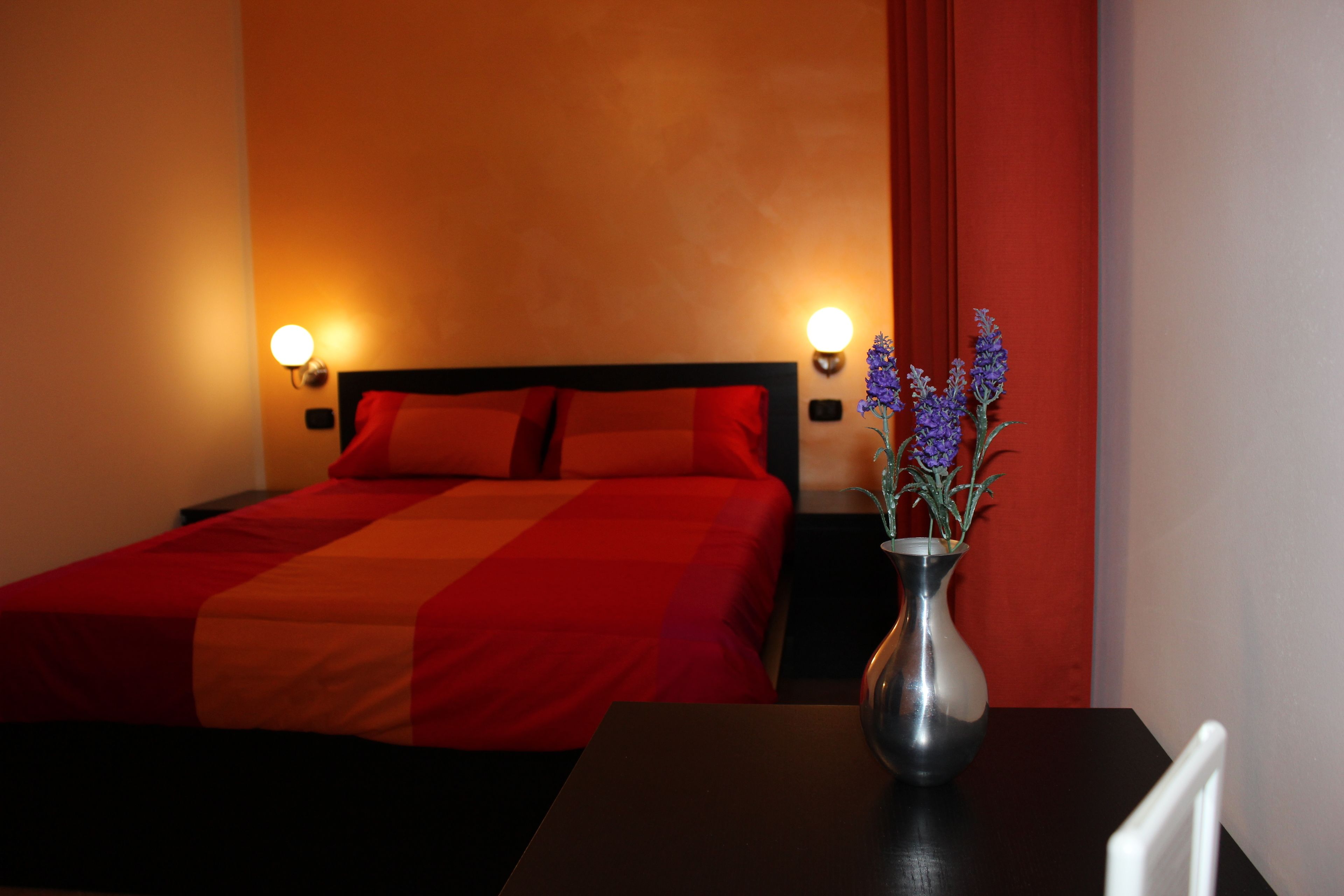 A hotel room with a bed, a nightstand, a vase with flowers, and wall sconces.