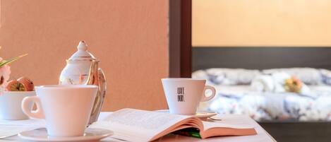 Una mesa ordenada con un libro, una tetera y dos tazas, una de ellas con la etiqueta 'Café Noir', en una habitación con una cama al fondo.