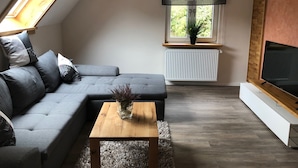 Ein modernes Wohnzimmer mit einem grauen Ecksofa, einem Holztisch, einem Fernseher auf einem Holzschrank, einer Heizung und einem Fenster mit Blick ins Grüne.