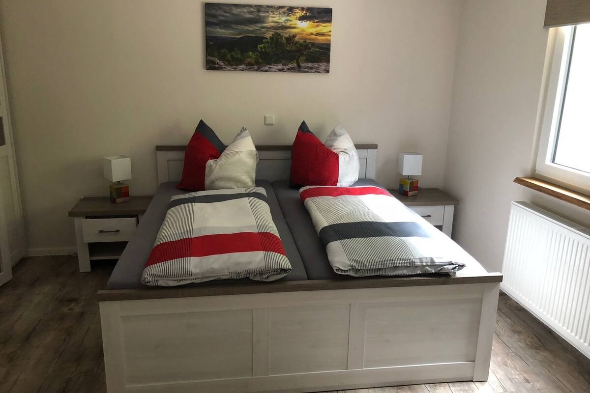 A double bed with a striped bedspread, two bedside tables with lamps, a nightstand, and a painting of a sunset on the wall.