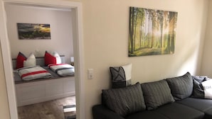 Ein Wohnzimmer mit einer schwarzen Couch, einer Bank mit Kissen, einem Waldgemälde und Holzboden.