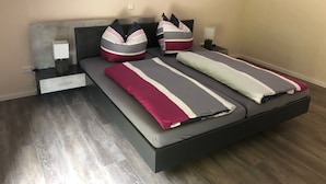 Ein modernes Schlafzimmer mit einer grauen Schlafcouch, einem Nachttisch mit Lampe und einem Beistelltisch ebenfalls mit Lampe.