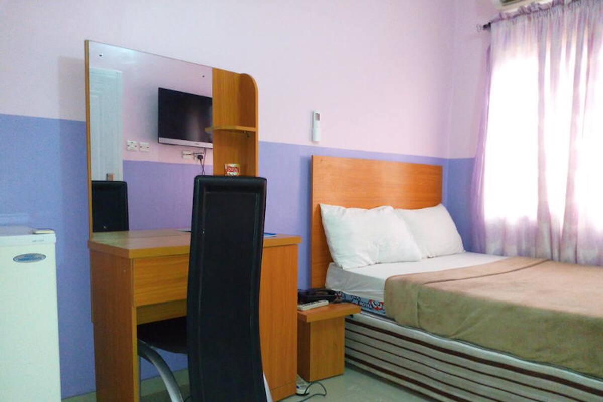 A hotel room with a wooden desk, a bed with a headboard, a television mounted on the wall, and a window with curtains.