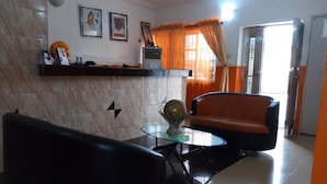 A living room with a black sofa, a glass coffee table, and a red-orange chair. There is a marble wall with framed pictures and a small shelf with decorative items. - Link Majestic B2 Suites and Hotel (Ibadan)