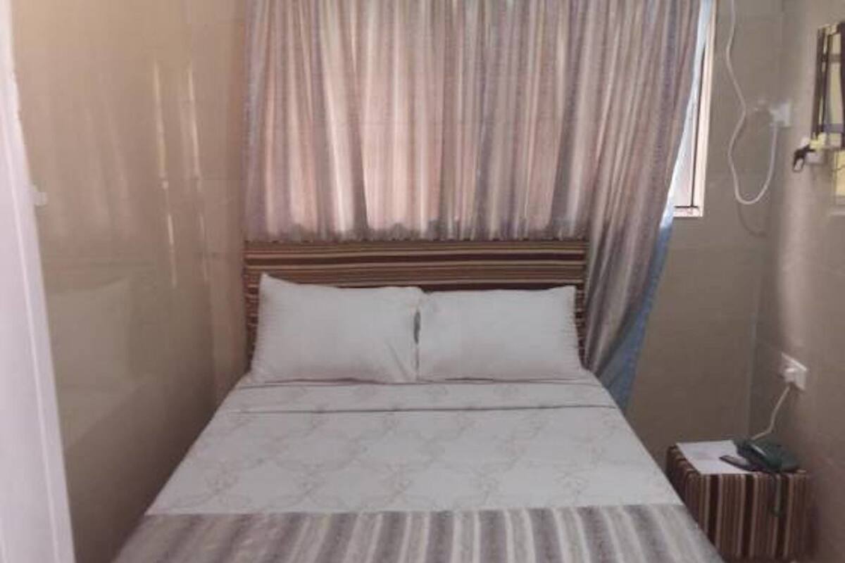 A single bed with a wooden headboard, a bedside table with a lamp, and a curtain.