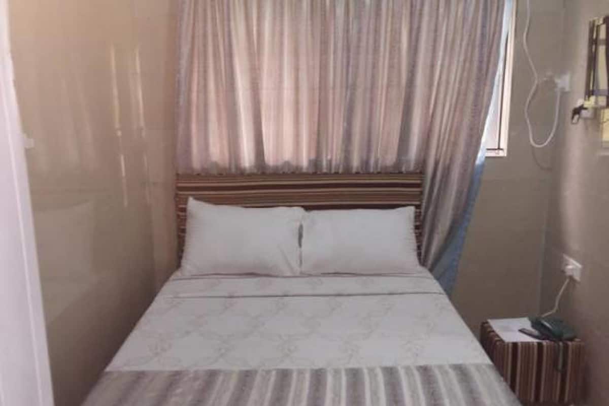 A single bed with a wooden headboard, a bedside table with a lamp, and a curtain.