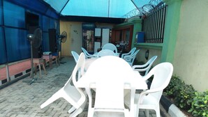 A covered outdoor seating area with white plastic chairs and tables, a Pepsi refrigerator, and a blue umbrella. - Link Majestic B2 Suites and Hotel (Ibadan)