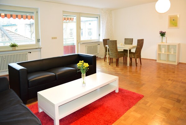 Een moderne woonkamer met een zwarte bank, een witte salontafel en een rood vloerkleed. Er is een eethoek met een tafel en stoelen, en een klein wit kastje met een plant.