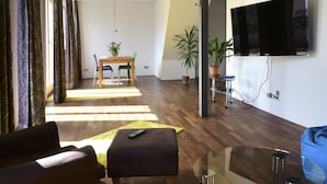 Un salon moderne avec un canapé, un pouf et une table basse en verre. Un téléviseur est fixé au mur, et il y a un coin repas avec une table et des chaises.