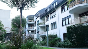 Un immeuble résidentiel à plusieurs étages, doté de balcons, d’un espace jardin et d’un chemin piétonnier dallé.
