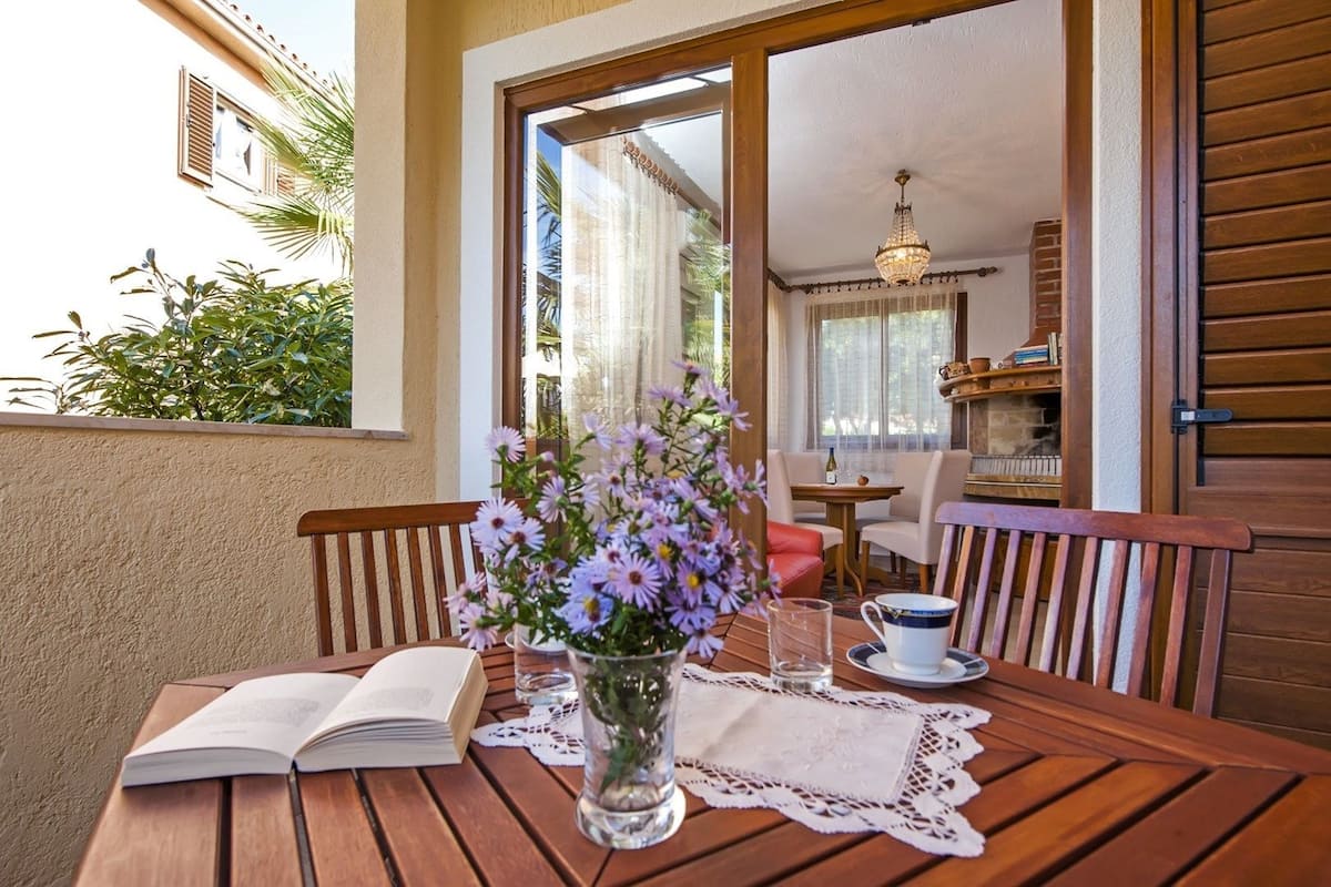 A wooden table with a book, a vase with flowers, and a glass of water on a patio.