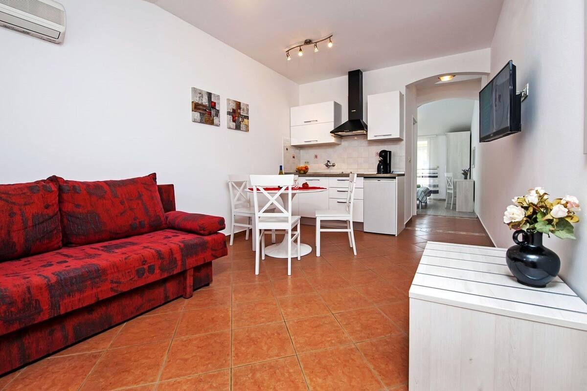 A compact living space with a red sofa, a dining table with chairs, a kitchen area, and a TV mounted on the wall.