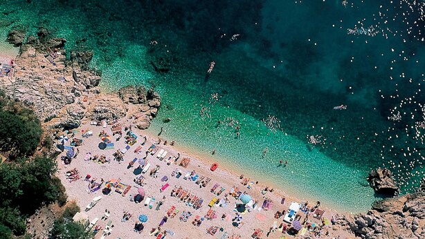 An aerial view of a beach with clear turquoise waters, rocky terrain, and numerous beach umbrellas and sun loungers. - Apartments Bastovanovic (Rovinj)