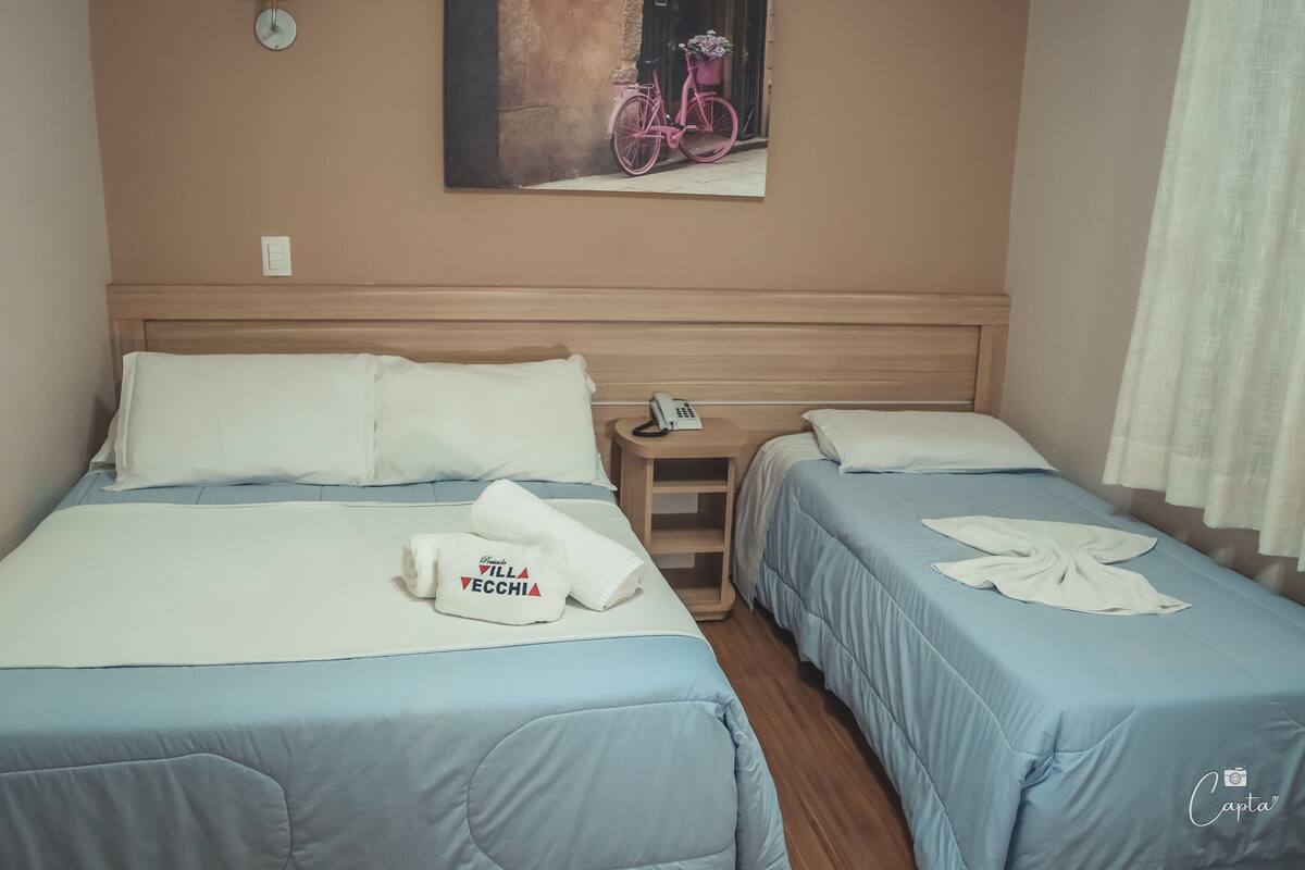 A hotel room with two beds, each with a blue bedspread and white pillows. There is a painting of a pink bicycle on the wall, a bedside table with a phone, and a towel with text on it.