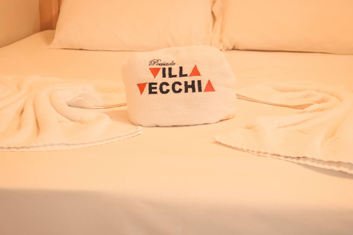 A bed with a white pillow and a white towel with the text "Pousada VILL ECCHI" on it.