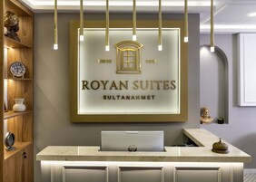 Una recepción con una computadora portátil, un cartel enmarcado que dice "ROYAN SUITES SULTANAHMET 2018" y un estante de madera con objetos decorativos.