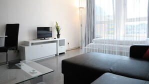 Een moderne woonkamer met een glazen salontafel, een zwarte bank, een witte tv-meubel en een groot raam met dunne gordijnen.
