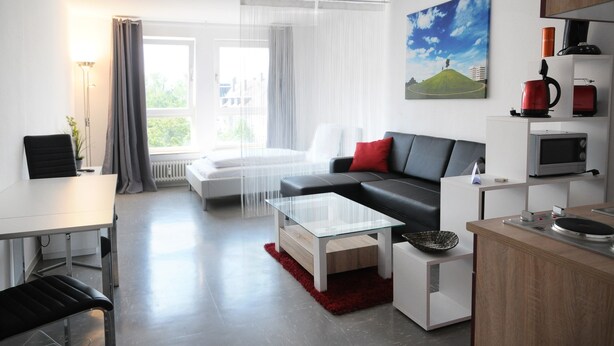 Een moderne woonkamer met een zwarte bank, een witte salontafel en een rood tapijt. Er is een eettafel met stoelen, een keuken met een magnetron, en een groot raam met dunne gordijnen.