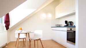 A modern kitchen with white cabinets, a built-in oven, and a coffee maker. There is a dining area with a table and two chairs.