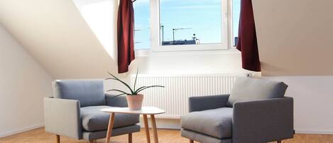 Una habitación moderna y luminosa con techo inclinado, dos sillones grises, una mesita redonda y una planta en maceta.