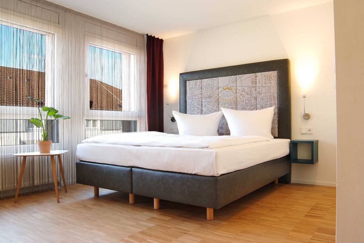 Une chambre d’hôtel moderne avec un grand lit, une tête de lit décorée d’une carte, une petite table avec une plante et une table de chevet.