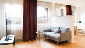 A modern hotel room with a sofa, a small table, and a view of the city through large windows.