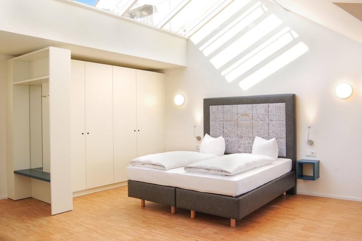 Une chambre avec une lucarne, un lit avec une literie blanche, une tête de lit grise et un mur décoré d’une carte.