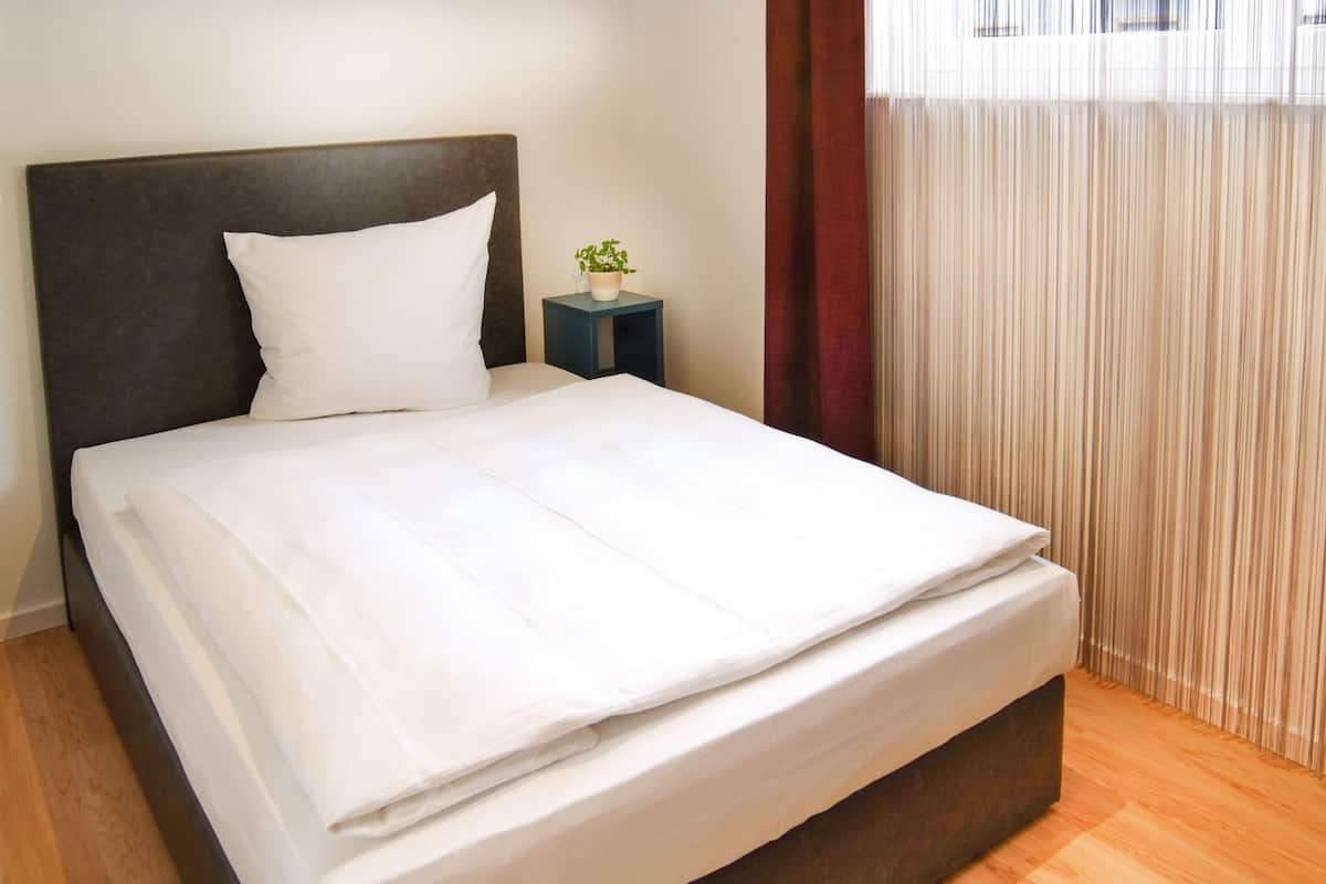 A neatly made bed with white linens in a room with wooden flooring and a dark headboard.