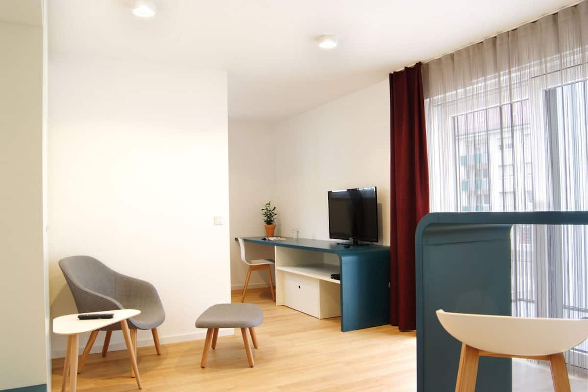 A modern, well-lit room with a wooden floor, a desk with a computer and TV, a grey chair, a small stool, and a window with sheer curtains.