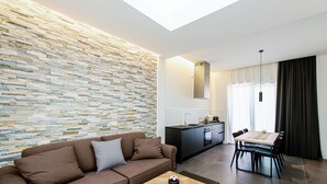 A modern living room with a stone wall, a brown sofa, a wooden coffee table, and a dining area with a table and chairs. - Acquarella Resort - Acquamarina Suite (TREVIGNANO ROMANO)