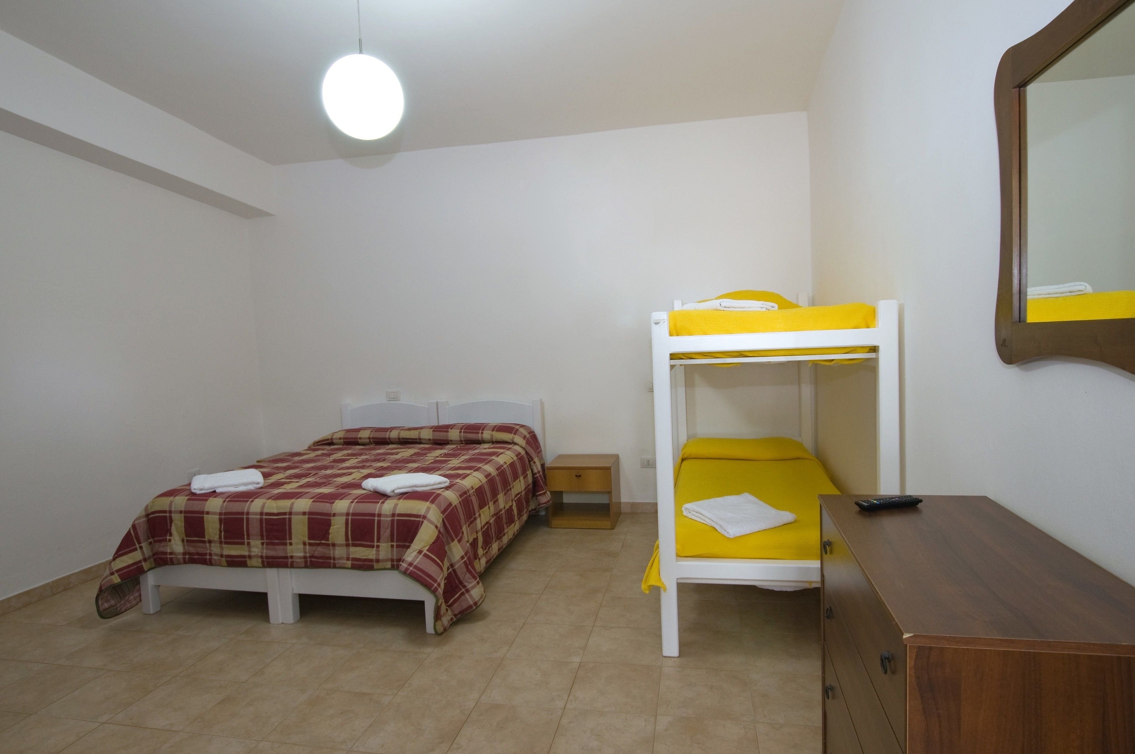 Una habitación con dos camas, una con colcha a cuadros roja y amarilla, y la otra con colcha amarilla. Hay un aparador de madera y un espejo en la pared.