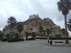 Ein historisches Gebäude mit einem markanten Turm auf einem Hügel, umgeben von Palmen und einer gepflasterten Fläche mit Bänken.