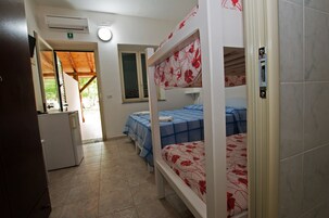 A dormitory room with bunk beds, a small kitchenette, and a window with a view of greenery outside.