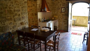 A rustic stone-walled dining area with a wooden table and chairs, a kitchen with a stove, and a doorway leading outside. - Agriturismo Marano (Gubbio)