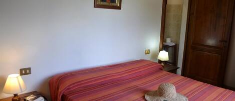 Un dormitorio con una colcha a rayas, una mesita de noche con una lámpara y un libro, un sombrero y un cuadro en la pared.