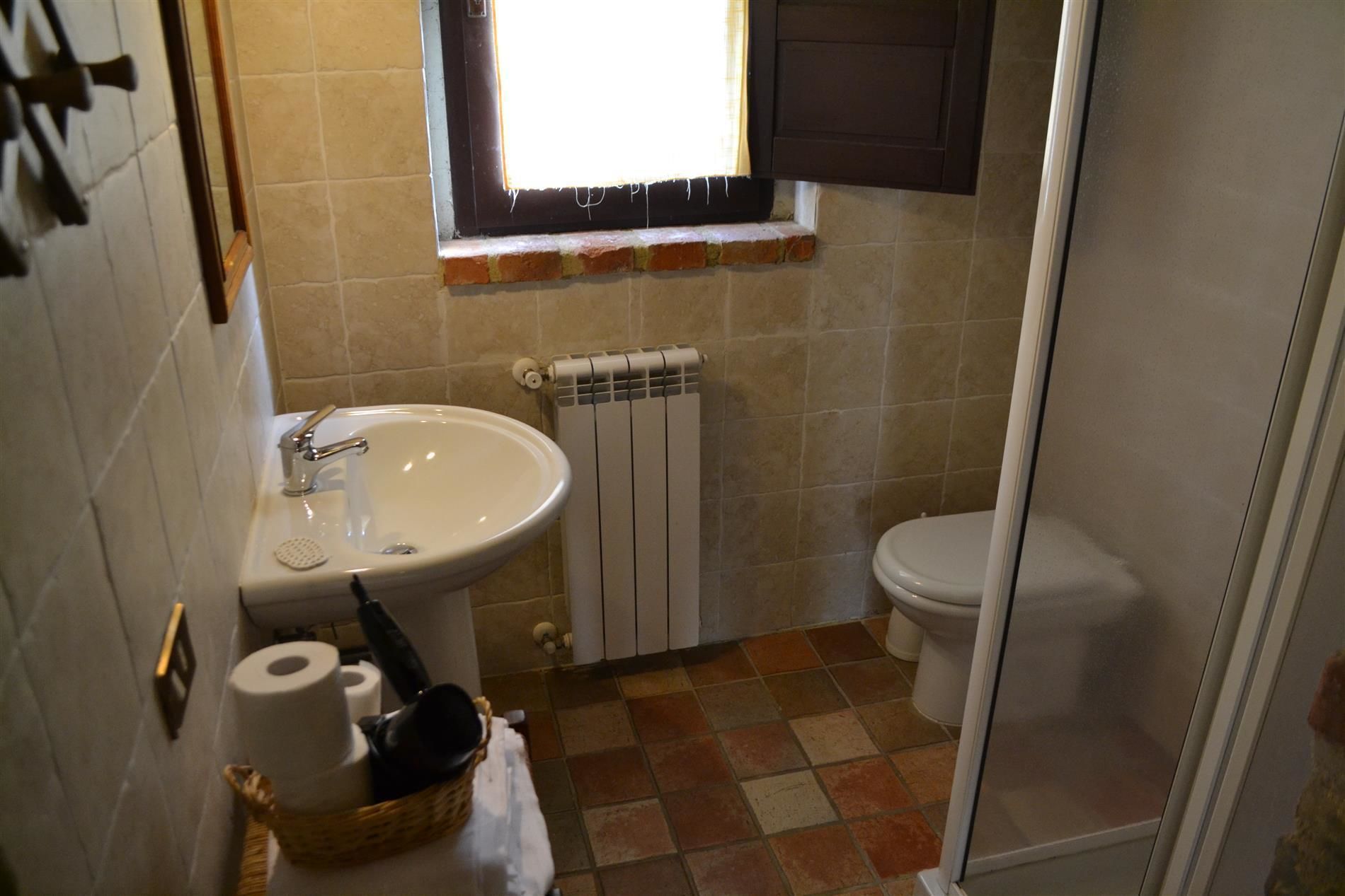 Baño con lavamanos blanco, inodoro y radiador. Hay una ventana sobre el lavamanos y una puerta de ducha a la derecha.
