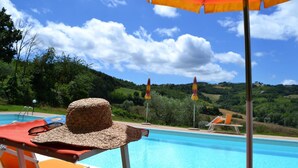 A poolside area with a hat on a chair, a pool, and a view of green hills under a clear sky. - Agriturismo Marano (Gubbio)
