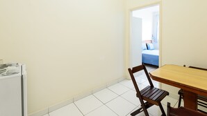 A small, clean room with a wooden dining table and chairs, a white tiled floor, and a view into another room with a bed and curtains. - Apartamento A1206 - Omar do Rio (Rio de Janeiro)