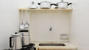 A compact kitchen with a gas stove, a sink, and storage shelves holding pots and pans. - Apartamento A1206 - Omar do Rio (Rio de Janeiro)