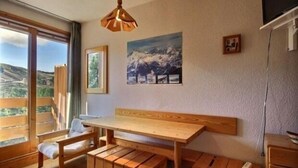 Una habitación con una mesa y un banco de madera, un cuadro de montañas y una ventana con vista a las colinas.